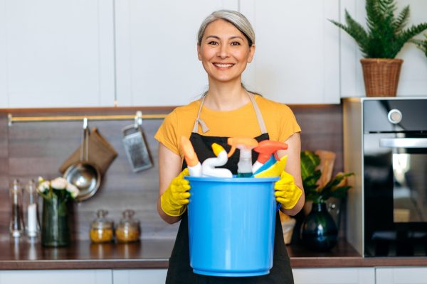 Beautiful senior gray-haired Asian housewife or cleaning lady in an apron and gloves stands in the kitchen, holds blue bucket with rags and household chemicals, looks at the camera, smiles