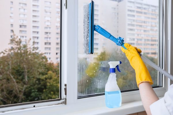 Man in yellow rubber gloves cleaning window with cleaner spray detergent and squeegee or rag at home or office, copy space. Housework and housekeeping, home hygiene, professional cleaning service